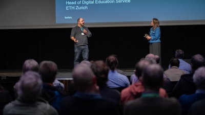 A presenter and an expert are on stage in front of the audience, presenting the day's highlights on a huge screen.